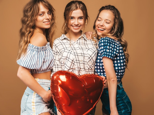 Drie glimlachende mooie vrouwen in de kleren van de geruite overhemdszomer. Meisjes poseren. Modellen met hartvormige ballon. Klaar voor Valentijnsdag van de viering