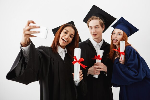 Drie internationale afgestudeerde vrienden die zich in mantels verheugen en een selfie op een telefoon maken. Toekomstige specialisten of medici hebben plezier met hun diploma's over de witte muur.