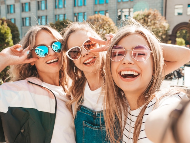 Drie jonge lachende hipster vrouwen in zomerkleren. Meisjes nemen selfie zelfportret foto's op smartphone. Modellen poseren in de straat. Vrouw met positieve gezicht emoties in zonnebril