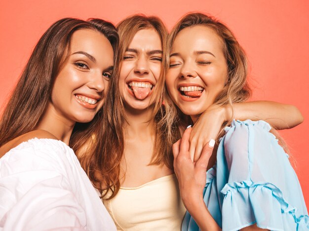 Drie jonge mooie glimlachende hipster meisjes in trendy zomerkleren. Sexy onbezorgde vrouwen die dichtbij roze muur stellen. Positieve modellen worden gek. Zelfportretfoto's maken op smartphone