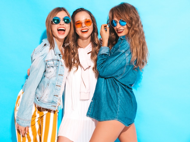 Drie jonge mooie glimlachende meisjes in trendy zomer kleurrijke kleding. Sexy onbezorgde die vrouwen in zonnebril op blauw worden geïsoleerd. Positieve modellen