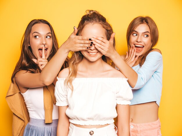 Drie jonge mooie lachende hipster meisjes in trendy zomerkleding. Sexy zorgeloze vrouwen poseren in de buurt van gele muur. Modellen verrassen hun vriend. Ze bedekken haar ogen en knuffelen van achteren