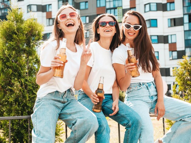 Drie jonge mooie lachende hipster vrouw in trendy zomer dezelfde kleren Sexy zorgeloze vrouwen poseren in de straat Positieve modellen plezier in zonnebril Drinkfles bier Oktoberfest