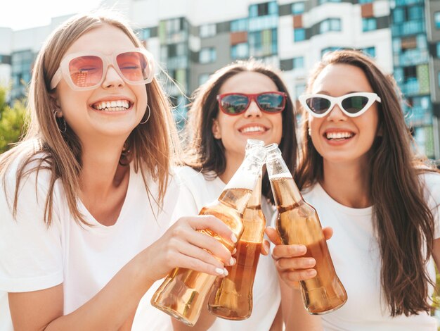 Drie jonge mooie lachende hipster vrouw in trendy zomer dezelfde kleren Sexy zorgeloze vrouwen poseren in de straat Positieve modellen plezier in zonnebril Drinkfles bier Oktoberfest