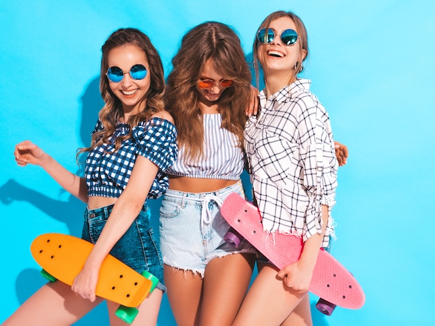 Drie jonge stijlvolle lachende mooie meisjes met kleurrijke penny skateboards. vrouw in de zomer geruit hemd kleren poseren. positieve modellen hebben plezier