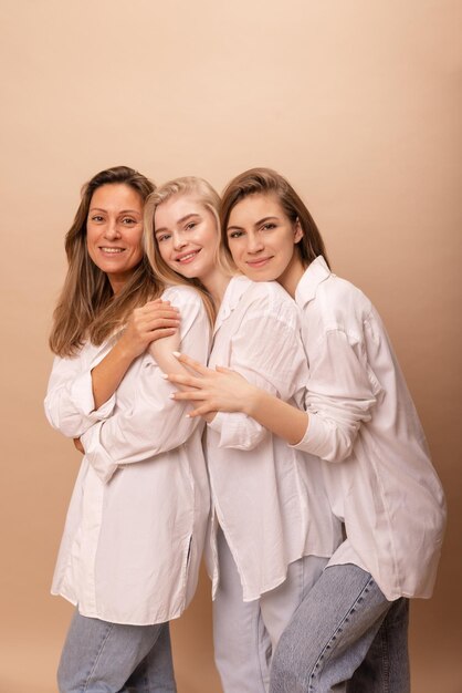 Drie jonge vrouwen met een lichte huid in witte overhemden glimlachen naar de camera en knuffelen elkaar tegen een beige achtergrond Concept van vrouwelijke schoonheid