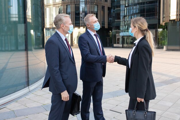 Drie kantoormedewerkers in gezichtsmaskers die deal of begroeting maken. Professionele succesvolle onderneemster en zakenlieden die zich buiten en handshaking bevinden. Onderhandeling, bescherming en partnerschap concept