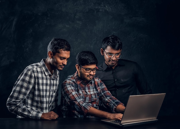 Drie kerels met een donkere huid die aan het chatten zijn in een laptop die zijwaarts aan een bureau zit in een studio op een zwarte achtergrond