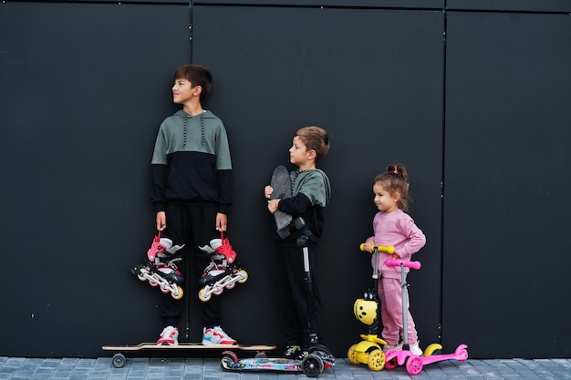 Drie kinderen buiten tegen zwarte moderne muur sportfamilie brengt vrije tijd buiten door met scooters en skates