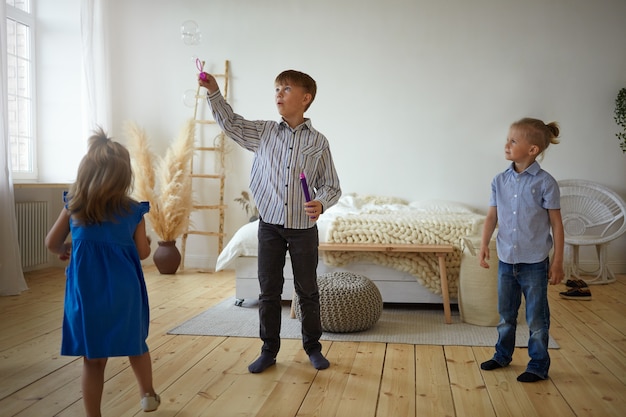 Drie kinderen spelen thuis samen. Schooljongen in shirt en spijkerbroek zeepbellen blazen in ruime slaapkamer, zijn kleine broer en zus wachten op hun beurt, staande op de vloer om hem heen