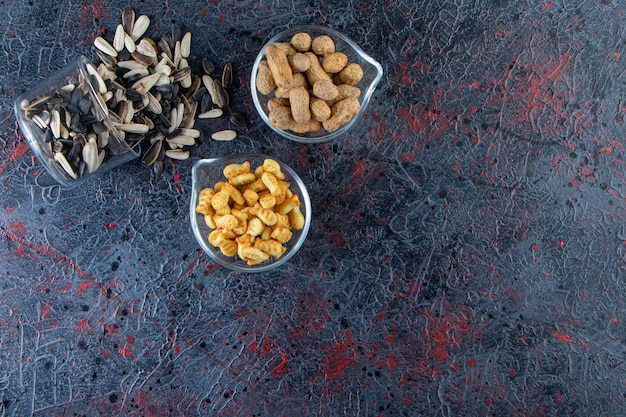 Drie kommen pinda's, zonnebloempitten en crackers op blauwe achtergrond.