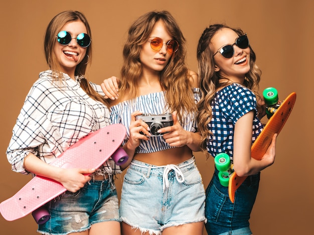 Drie mooie stijlvolle lachende meisjes met kleurrijke penny skateboards. vrouwen in zomer geruite shirt kleding poseren. foto's maken met een retro fotocamera