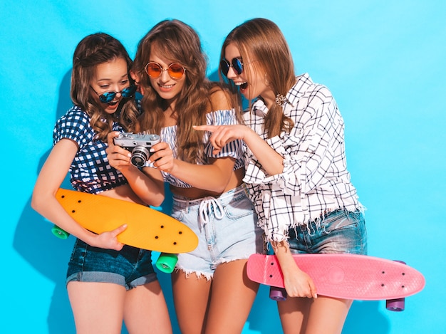 Drie mooie stijlvolle lachende meisjes met penny skateboards. Vrouwen in de zomer geruite shirt kleding en zonnebril. Foto's maken met een retro fotocamera