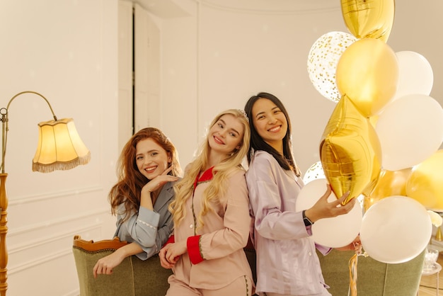 Drie prachtige jonge meisjes tussen verschillende rassen in zijden pyjama's vieren verjaardag in kamer met ballonnen Concept van speelse stemmingsvakantie