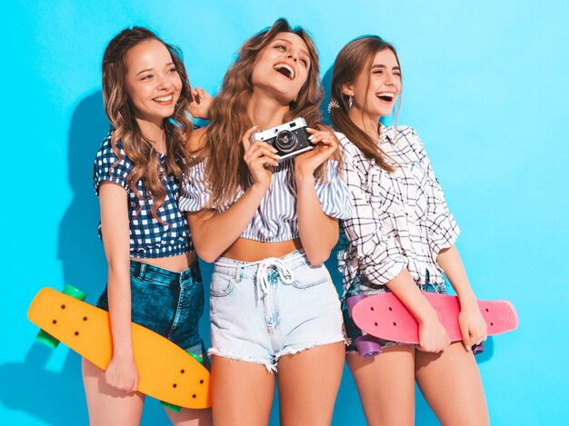 Drie sexy mooie stijlvolle lachende meisjes met kleurrijke penny skateboards. Vrouwen in zomer geruite shirt kleding poseren. Modellen die foto's maken op retro fotocamera