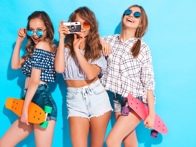 Drie sexy mooie stijlvolle lachende meisjes met kleurrijke penny skateboards. Vrouwen in zomer geruite shirt kleding poseren. Modellen die foto's maken op retro fotocamera