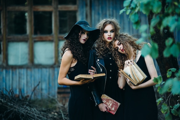 Drie vintage vrouwen als heksen poseren voor een verlaten gebouw met boeken in de hand aan de vooravond van halloween