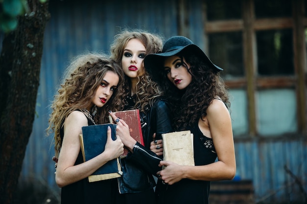 Drie vintage vrouwen als heksen poseren voor een verlaten gebouw met boeken in de hand aan de vooravond van Halloween