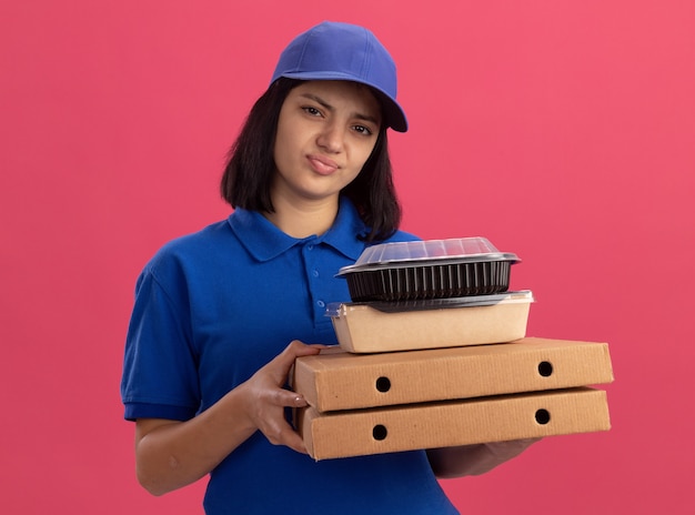 Droevig jong bezorgmeisje in blauw uniform en pet met pizzadozen en voedselpakket op zoek ontevreden staande over roze muur