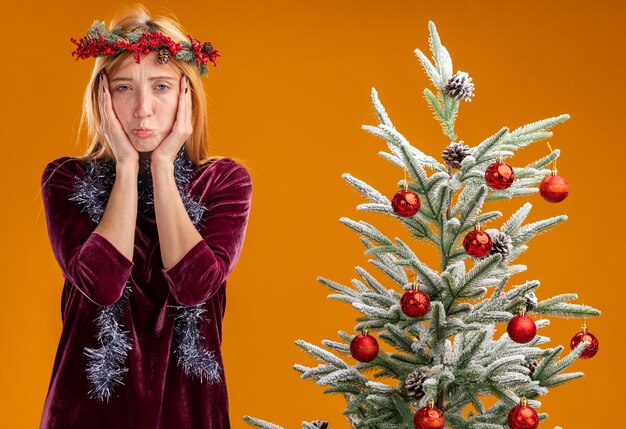 Droevig jong mooi meisje dat in de buurt van de kerstboom staat met een rode jurk en een krans met een krans op een nek bedekt gezicht met handen geïsoleerd op een oranje muur
