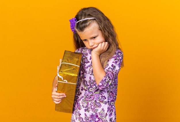 droevig klein blond meisje dat de hand op de kin legt en een geschenkdoos vasthoudt die op een oranje muur is geïsoleerd met kopieerruimte