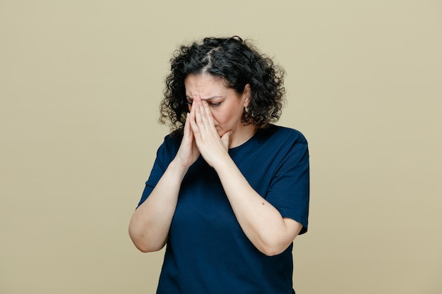 droevige en angstige vrouw van middelbare leeftijd die een t-shirt draagt, de handen bij elkaar houdt op de neus en neerkijkt op een olijfgroene achtergrond