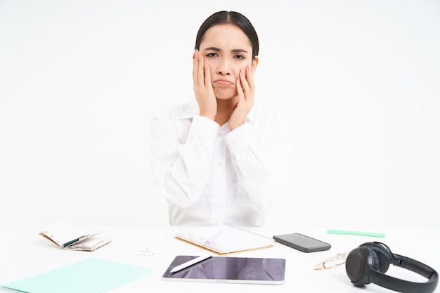 Gratis foto droevige en knorrige aziatische vrouw op de werkplek zit met digitale tablet en werkdocumenten zien er boos uit en