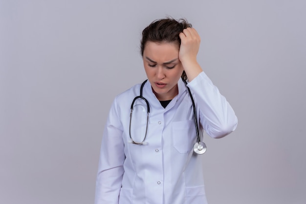 Droevige jonge arts die medische jurk draagt die een stethoscoop draagt, grijpt het hoofd op een witte muur