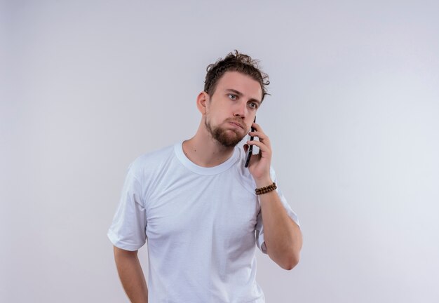 Droevige jonge kerel die een wit t-shirt draagt, spreekt over telefoon op geïsoleerde witte achtergrond