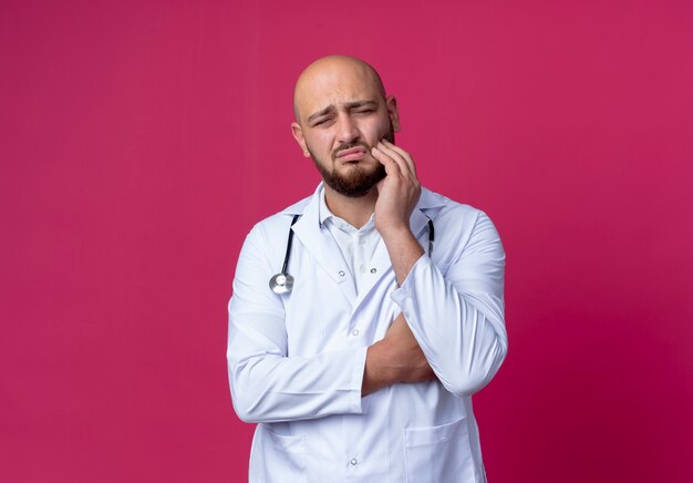 Droevige jonge mannelijke arts die medische mantel en stethoscoop draagt die hand op pijnlijke tand zet die op roze wordt geïsoleerd
