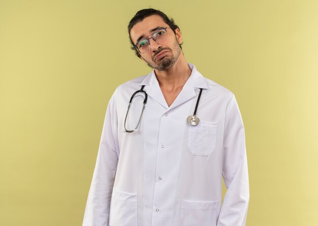 Droevige jonge mannelijke arts met optische bril die wit gewaad met stethoscoop op geïsoleerde groene muur met exemplaarruimte draagt