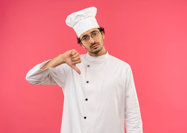 Droevige jonge mannelijke kok die eenvormige chef-kok en glazen zijn duim draagt die neer op roze muur wordt geïsoleerd