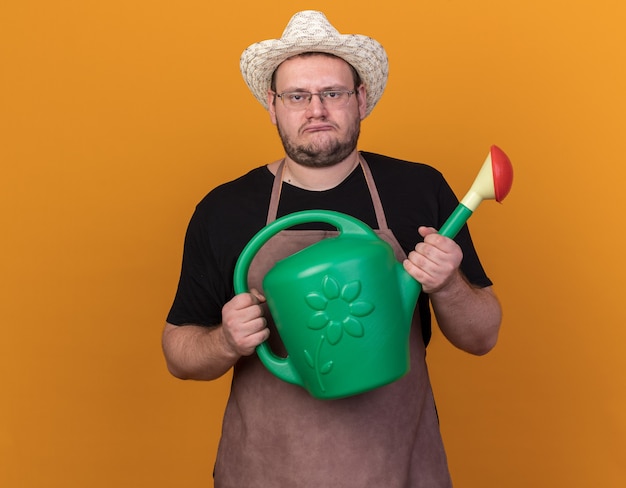 Droevige jonge mannelijke tuinman die tuinieren hoed draagt die gieter houdt die op oranje muur wordt geïsoleerd