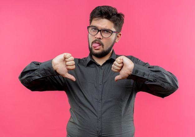 Droevige jonge zakenman die een bril draagt die tong en zijn duim toont neer geïsoleerd op roze