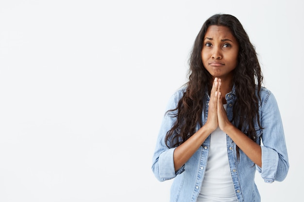 Droevige ongelukkige Afro-Amerikaanse vrouw die een spijkerblouse draagt, kijkend naar de ogen vol hoop en geloof tijdens het bidden, om hulp of vergeving vraagt, de handpalmen tegen elkaar gedrukt, poserend op wit