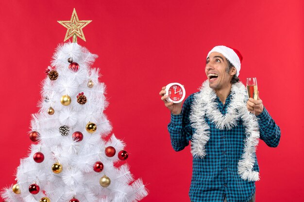 Dromer jonge man met kerstman hoed en met een glas wijn en klok staande in de buurt van de kerstboom op rood