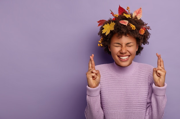 Dromerig hoopvol Afrikaans Amerikaans meisje gelooft in overwinning, bidt in de herfst, draagt gebreide trui, heeft bladeren in krullend haar geïsoleerd over violette muur