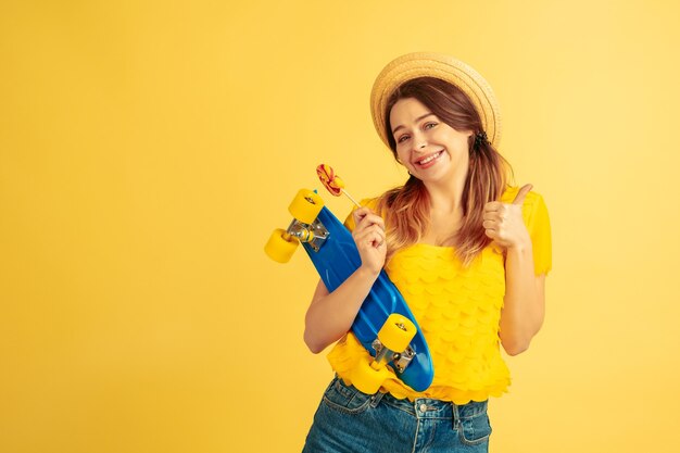 Dromerig, houdt skate vast. Het portret van de Kaukasische vrouw op gele studioachtergrond. Mooi vrouwelijk model in hoed. Concept van menselijke emoties, gezichtsuitdrukking, verkoop, advertentie. Zomer, reizen, toevlucht.