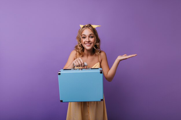 Dromerig krullend meisje in gele outfit poseren met bagage. Portret van debonair vrouwelijk model met koffer die naar camera glimlacht.