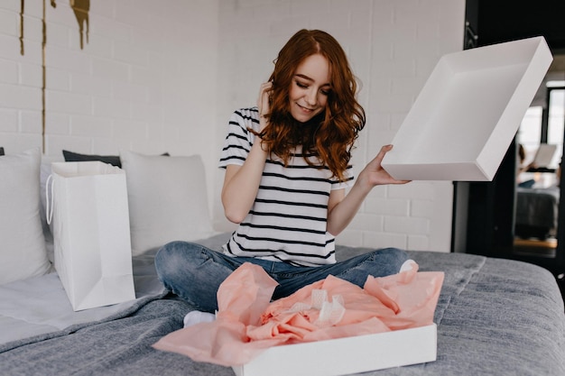 Dromerig meisje in gestreept t-shirt zittend op bed Extatische roodharige krullende vrouw poseren in slaapkamer