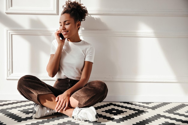 Dromerige Afro-Amerikaanse dame praten op smartphone Elegante zwarte meid zittend op de vloer onder zonnestralen