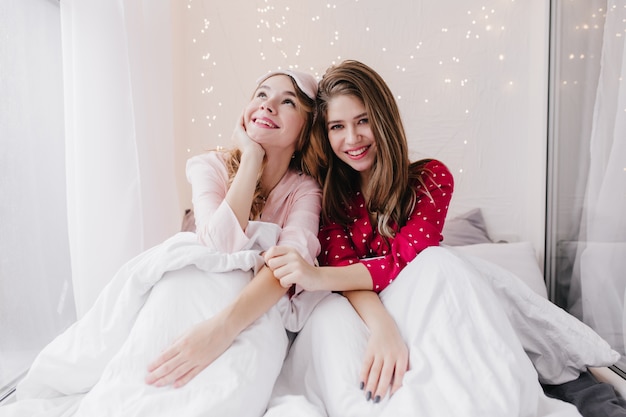 Dromerige blanke meisjes poseren onder een deken met een geïnspireerde glimlach. Indoor portret van aantrekkelijke Europese dames draagt pyjama's ontspannen in de ochtend.