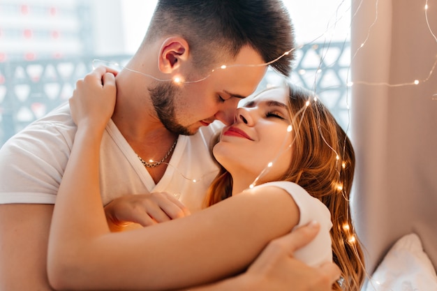 Dromerige blonde vrouw die haar donkerharige echtgenoot zachtjes omhelst. Knap mannelijk model kussend blond meisje.