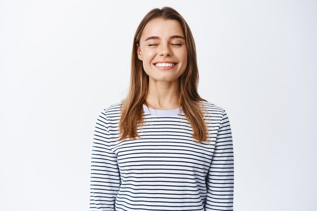 Dromerige en gelukkige mooie vrouw, ogen sluiten en glimlachen, vrij ademen, dagdromen of iets positiefs voorstellen, in casual outfit over witte muur staan