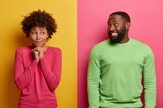 Dromerige etnische vrouw houdt de handen onder de kin, kijkt opzij, draagt een roze trui, een blije bebaarde man kijkt naar zijn vriendin, glimlacht gelukkig. mensen drukken positieve emoties uit, staan samen binnen.