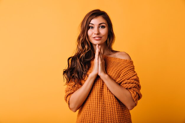 Dromerige jonge dame in een stijlvolle oranje trui poseren met een schattige glimlach. Indoor portret van schattig Kaukasisch meisje met lang golvend haar.