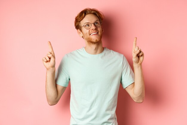 Dromerige jonge man met rood haar en baard die met de vingers wijst en vrolijk omhoog kijkt, bewondert iets...