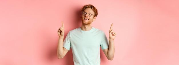 Gratis foto dromerige jonge man met rood haar en baard die met de vingers wijst en vrolijk opkijkt bewondert iets