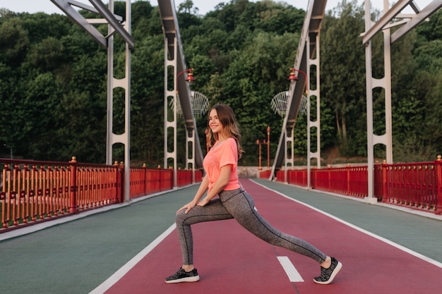 Dromerige krullende vrouw in sportbroek die zich uitstrekt op sintelpad. Openluchtportret van romantische meisjestraining