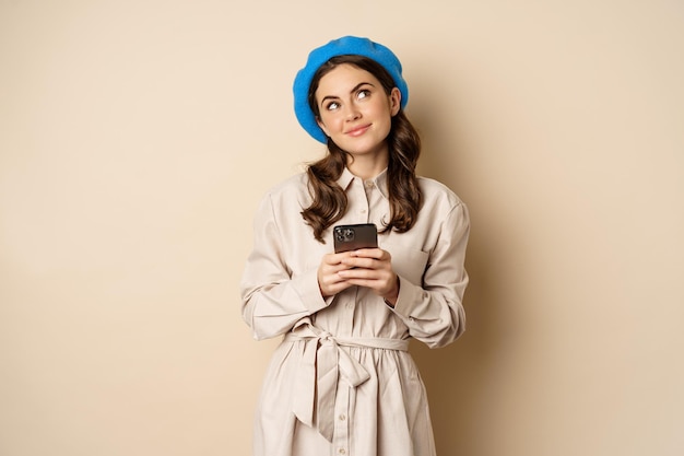 Dromerige lachende vrouw in stijlvolle trenchcoat, fantaserend opkijkend tijdens het winkelen op de mobiele telefoon-app, smartphone gebruikend en denkend, staande over een beige achtergrond.